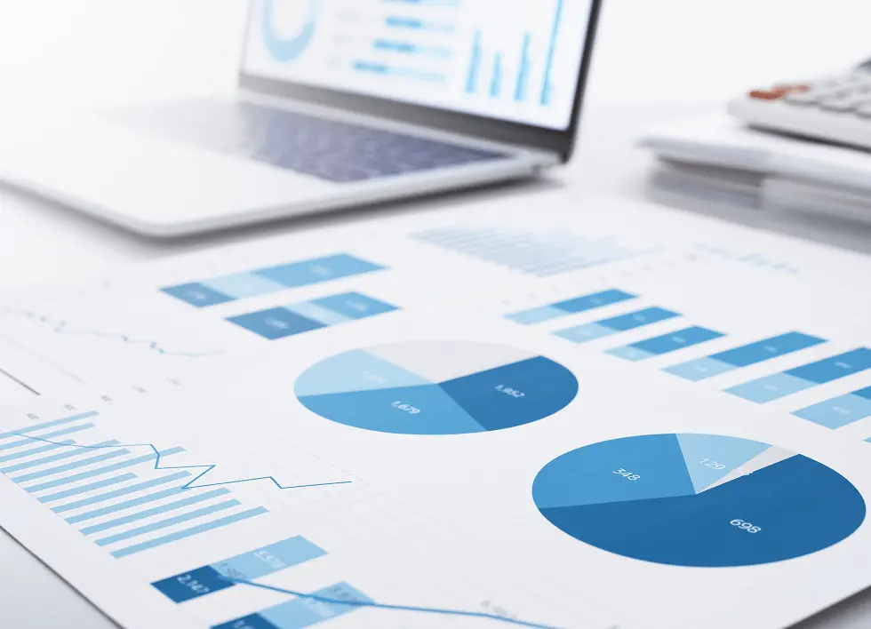デスク上に広げられた青色のグラフや円グラフが印刷されたビジネス資料。背景にはノートパソコンの画面に同様のグラフが表示されており、電卓も置かれている