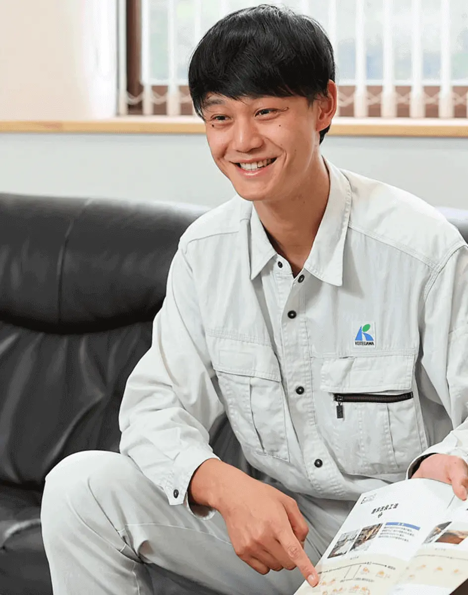 作業着の社員がソファに座り、資料を指し示しながら笑顔で説明している様子