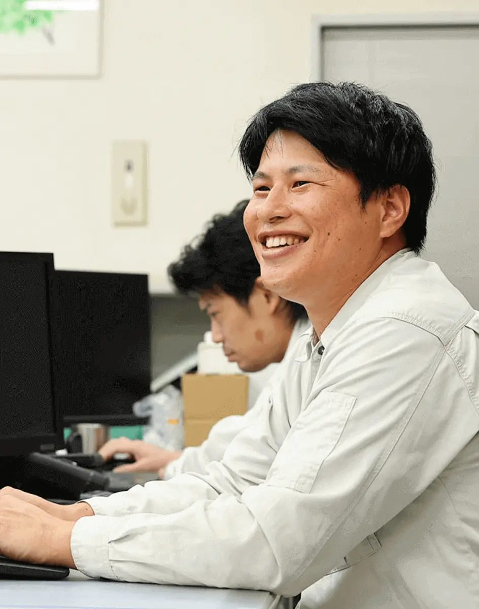 作業服の社員がデスクでパソコンを操作しながら笑顔で業務を行っている様子