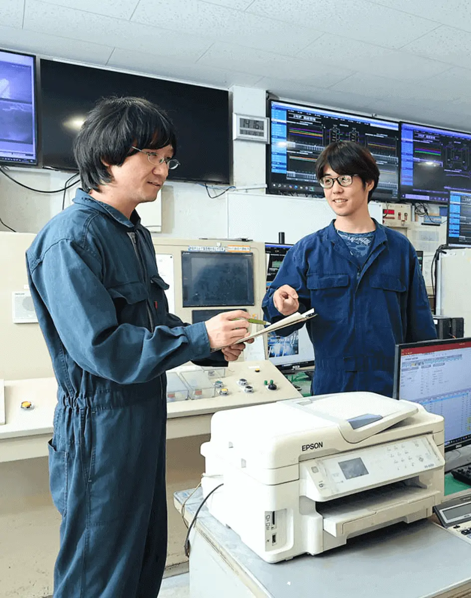 工場の制御室で二人の作業員が計器やモニターを確認しながら機器について話し合っている様子