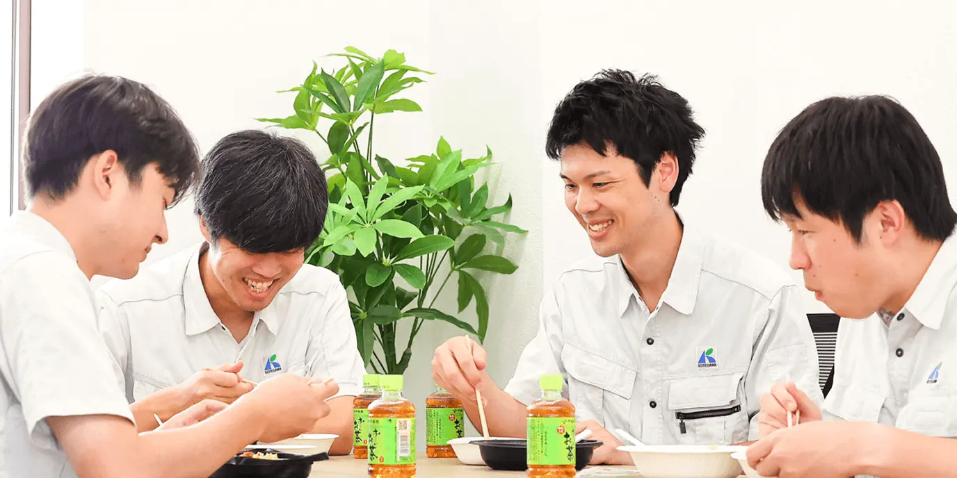 作業着姿の社員4人が食事を囲んで笑い合う様子。机の上にはお茶のボトルや弁当が並び、和やかな雰囲気が伝わる。