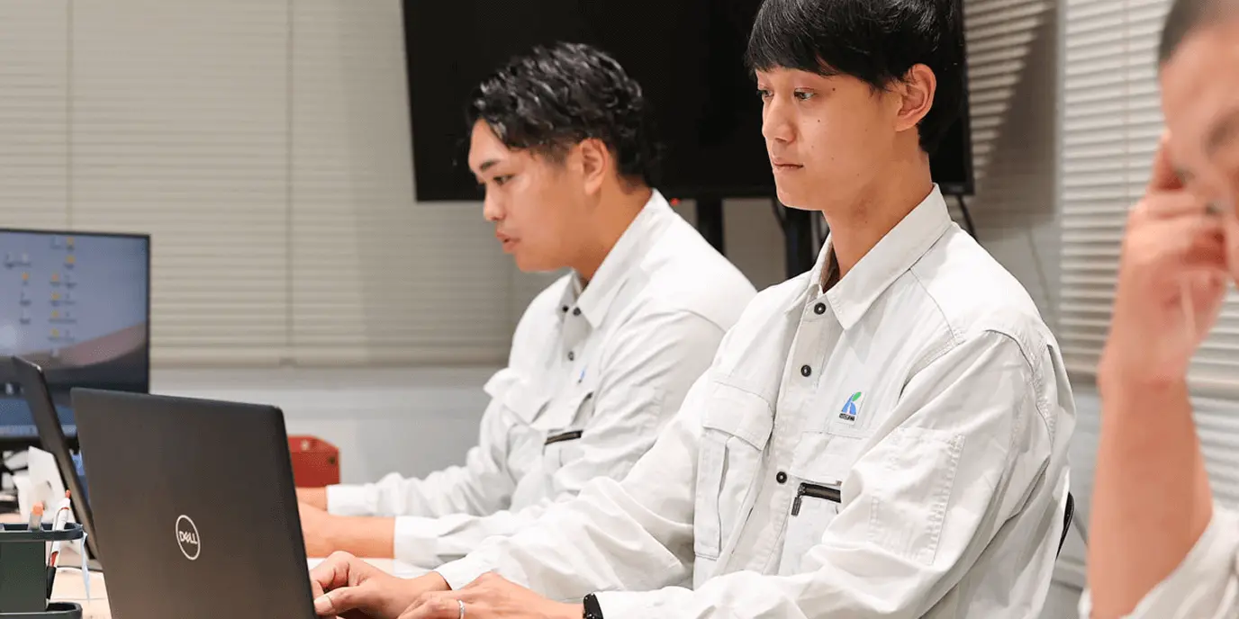 作業着を着た社員がパソコンに向かい集中して作業している様子。オフィス内の落ち着いた作業風景。