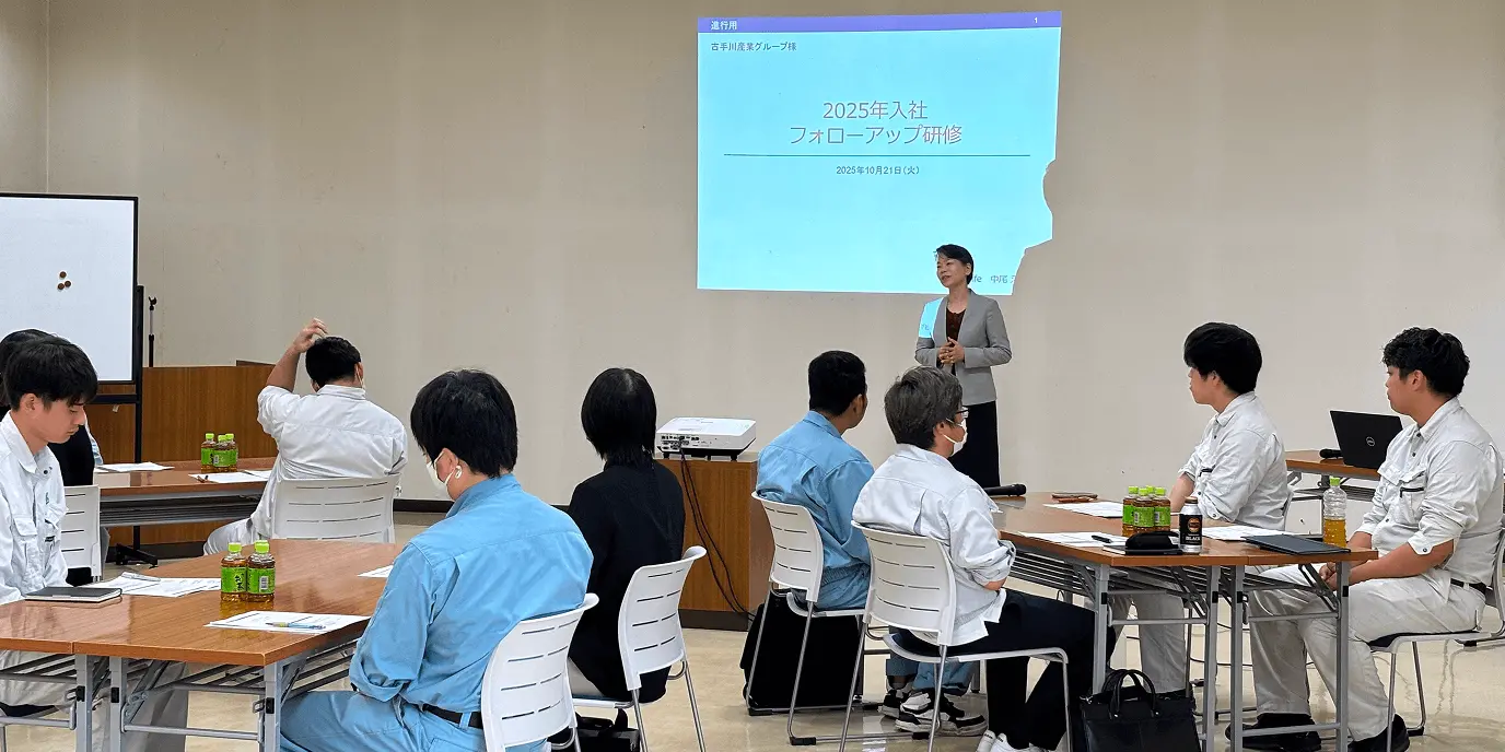 研修室で講師が前に立ち、スクリーンに「2025年入社フォローアップ研修」と表示された資料を映して説明している。複数の参加者がテーブルに着席し、講義を受けている様子。