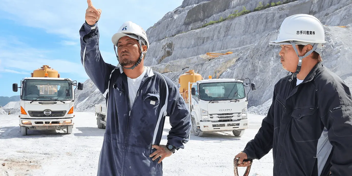 ヘルメットを着用した作業員が採石場で指示を出している様子。背景には大型トラックと岩場が広がる。