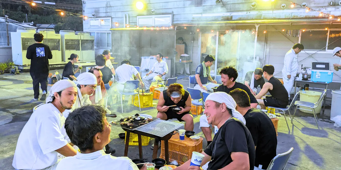 屋外で従業員たちが集まり、バーベキューを楽しみながら談笑している様子。夜の明かりの下でリラックスした雰囲気が伝わる。