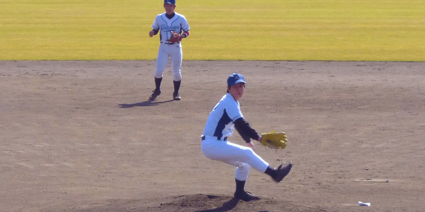 野球の試合で、ピッチャーマウンドに立つ選手が投球動作を行っている様子。後方には守備位置についた選手が見える。