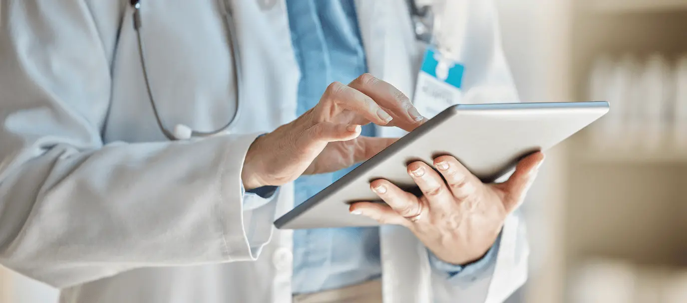 タブレット端末を操作しながらデジタル記録を確認する医療従事者の手元。白衣と聴診器が写り、業務でのデジタル活用の様子が伝わる場面。