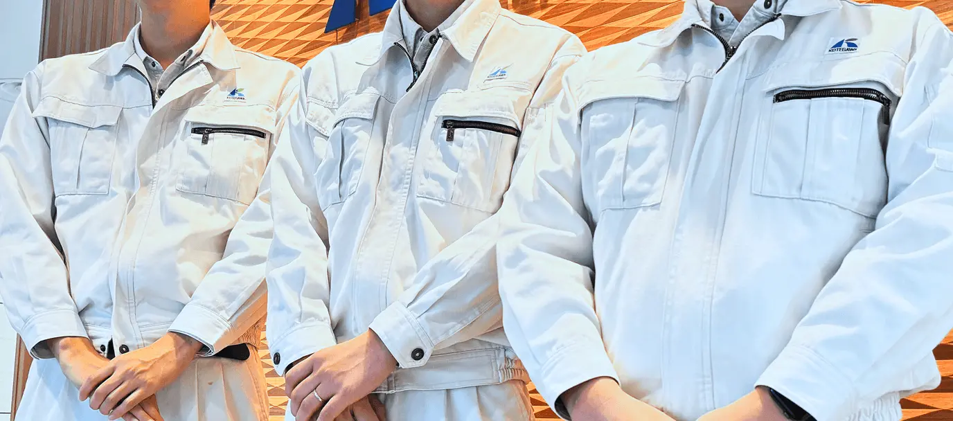 作業服を着た3人の社員が並んで立っている様子。胸元には古手川産業のロゴが入り、明るいエントランス前で撮影された集合カット。