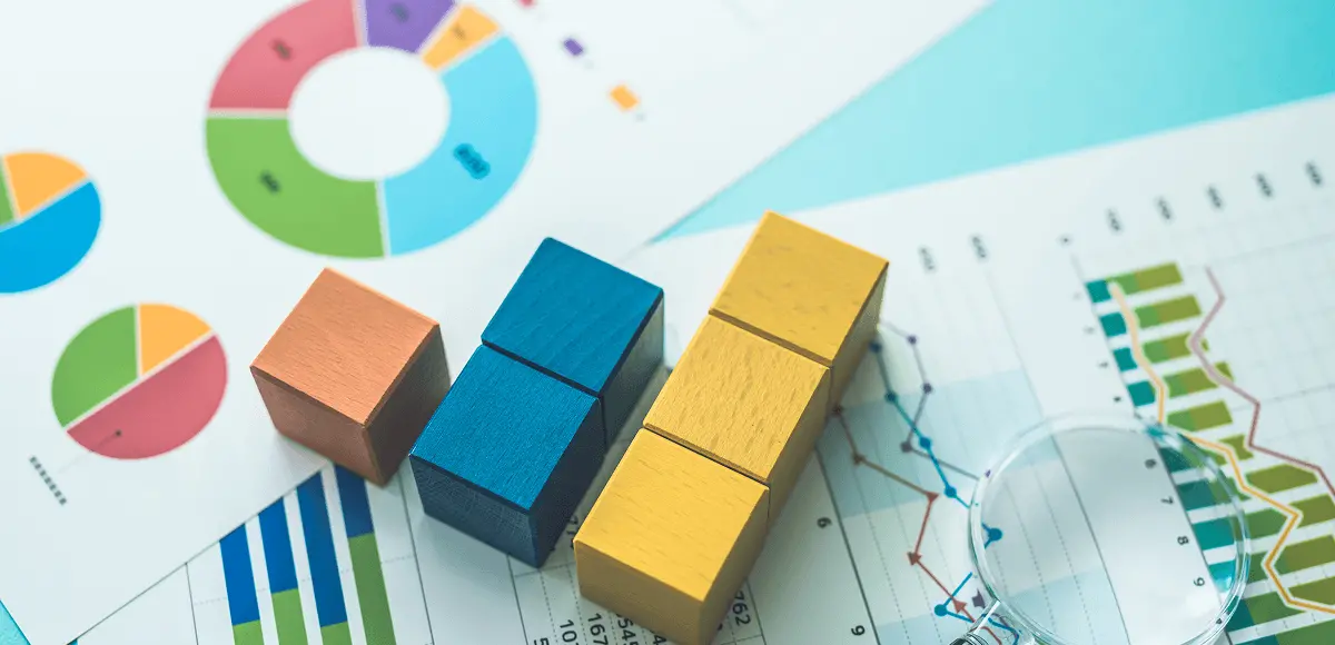 円グラフや棒グラフが印刷された資料の上に、色の異なる木製キューブブロックが並べられ、横には虫眼鏡が置かれている。データ分析や統計の概念を表す構図。
