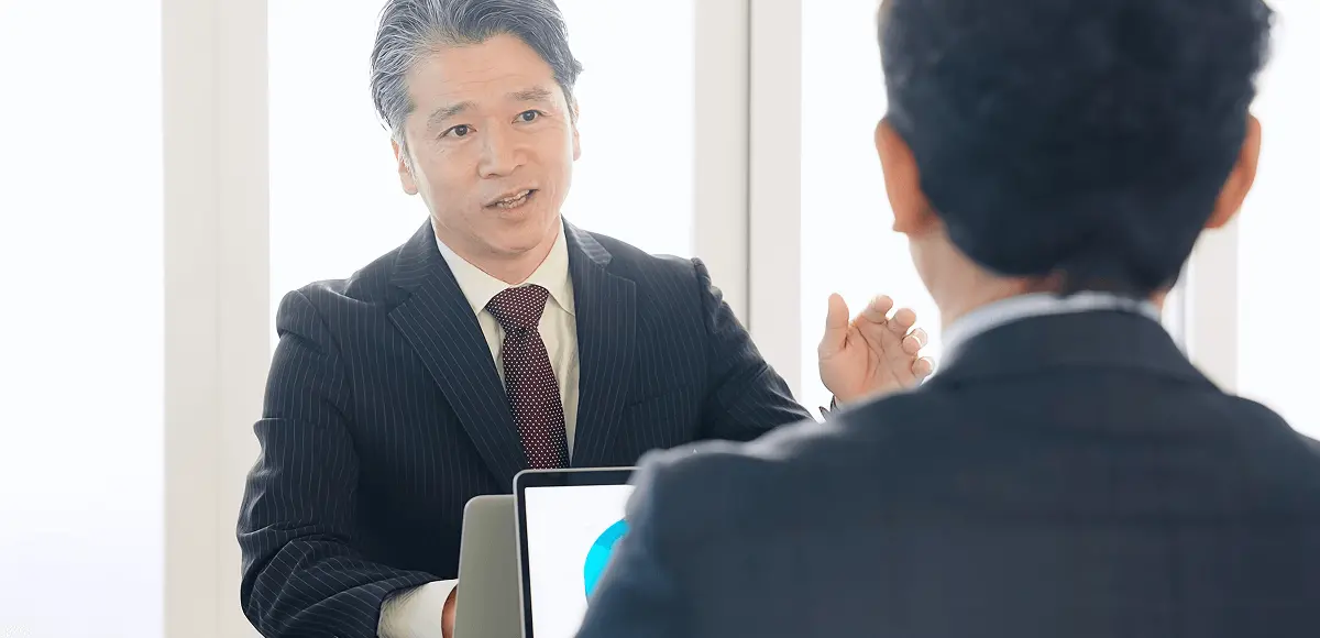 スーツ姿のビジネスマン2人が向かい合い、1人がノートパソコンの画面を示しながら説明している様子。ビジネス会議や面談の場面を表している。