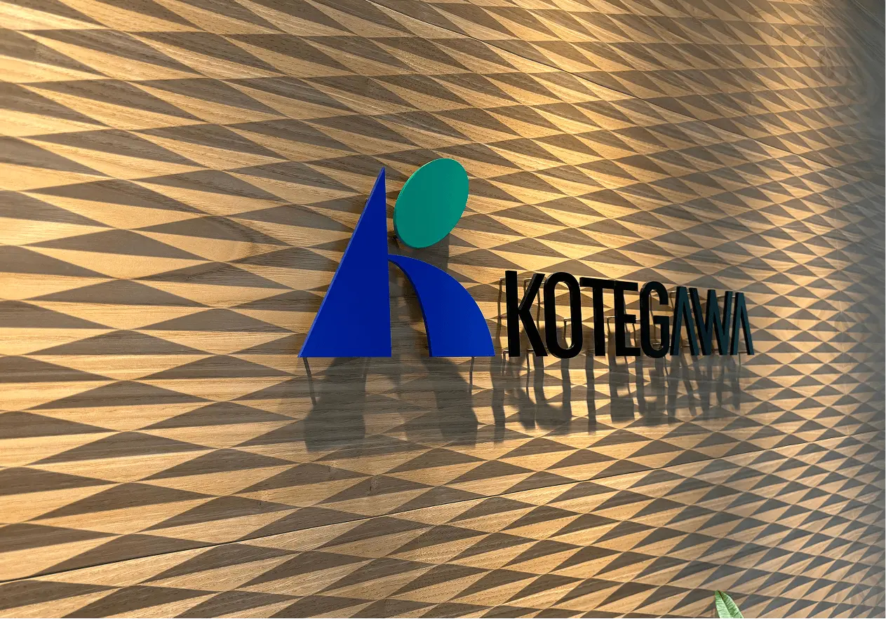 木目調の壁に掲げられた古手川産業のロゴマークと「KOTEGAWA」の文字。立体的なロゴが照明により陰影を帯びて浮かび上がっている。