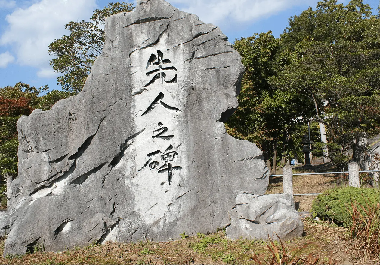 屋外に設置された大きな石碑で、「先人之碑」と刻まれている。背景には樹木と青空が広がっている。