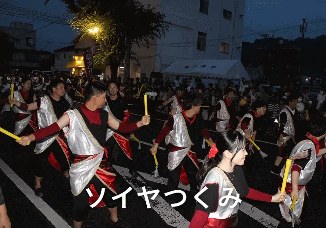 夜の街中で、白と赤と黒の衣装を着た踊り手たちが黄色い鳴り物を持って列を成して踊っている、ソイヤつくみ祭りの様子。背景には多くの観客と白いテントが見える