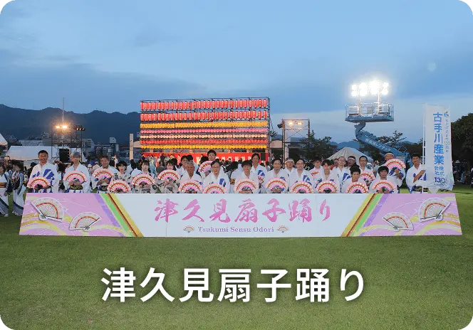 夕暮れ時の屋外イベント会場で、白い法被姿の参加者たちが扇子を持って集合写真を撮影している様子。前方には「津久見扇子踊り」と書かれたピンク色の横断幕が掲げられ、背景には提灯で飾られた櫓が見える津久見扇子踊りの風景