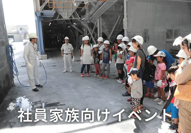 工場内の作業場で、白いヘルメットをかぶった子どもたちと保護者が、作業服姿の従業員から施設の説明を受けている社員家族向け工場見学イベントの様子