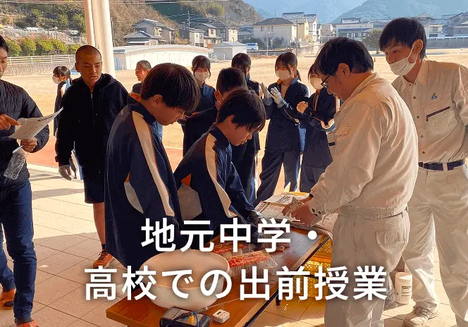 屋外のテラスで、制服姿の中学生や高校生が、作業服を着た講師から木材を使った実習指導を受けている地元中学・高校での出前授業の風景