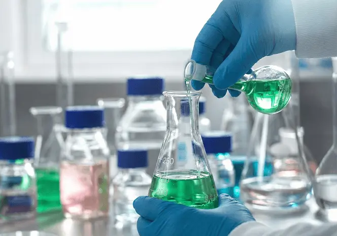 研究室で青い手袋をした研究者が、緑色の液体が入ったフラスコを慎重に持ち上げて観察している様子。周囲には試薬瓶やビーカーが並び、化学実験の場面を示している。