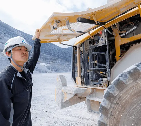 白いヘルメットをかぶった作業服姿の男性が、採石場で青いリモコン装置を操作している様子。背景には重機やトラックが見え、晴れた空の下での石灰石採掘作業の風景