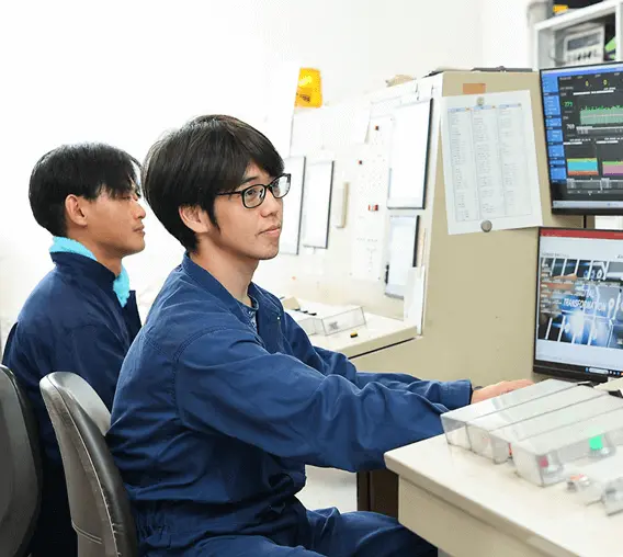 管理室内で、紺色の作業服を着た2名の若手社員が並んで座り、モニター画面を見ながら製造プロセスの監視・制御業務を行っている様子。背景には図面や資料が掲示されている