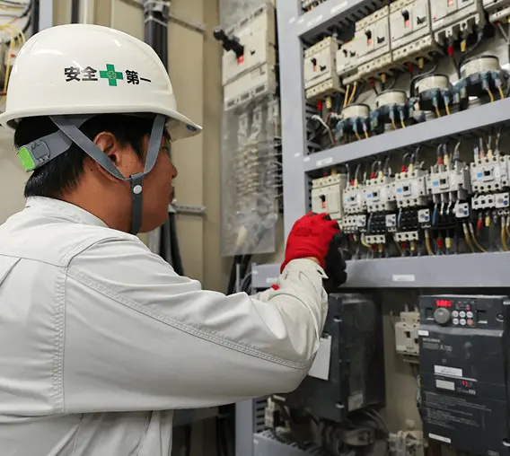 配電盤の前で作業員がヘルメットと手袋を着用し、電気設備の点検作業を行っている様子。多数の配線やスイッチが並ぶ制御盤を慎重に操作している。