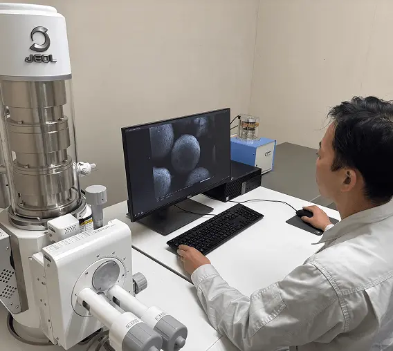 分析装置の前で作業員がコンピューター画面に表示された粒子の拡大画像を確認している様子。電子顕微鏡を用いて材料の表面状態を観察・分析している場面。
