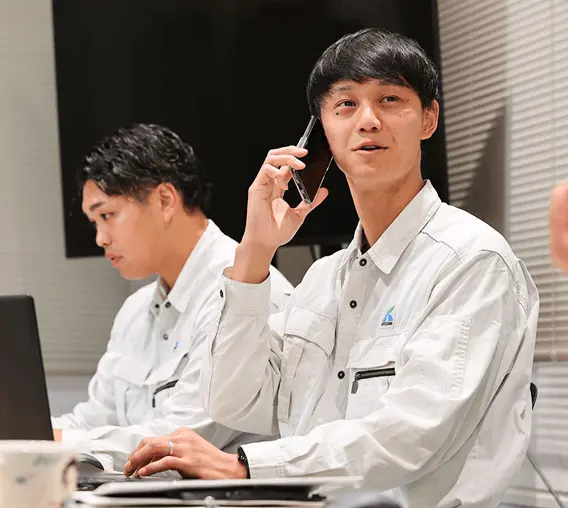 オフィス内で、白い作業服を着た2名の社員が携帯電話で通話したりパソコン作業を行っている。手前の社員は立って電話をしており、奥の社員は着席してデスクワークをしている業務風景