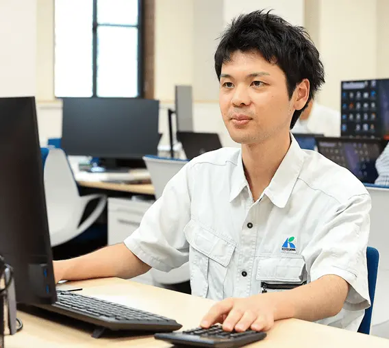 オフィスでパソコンを操作する社員の男性。作業服を着用し、明るい職場環境の中でデスクワークを行っている様子。