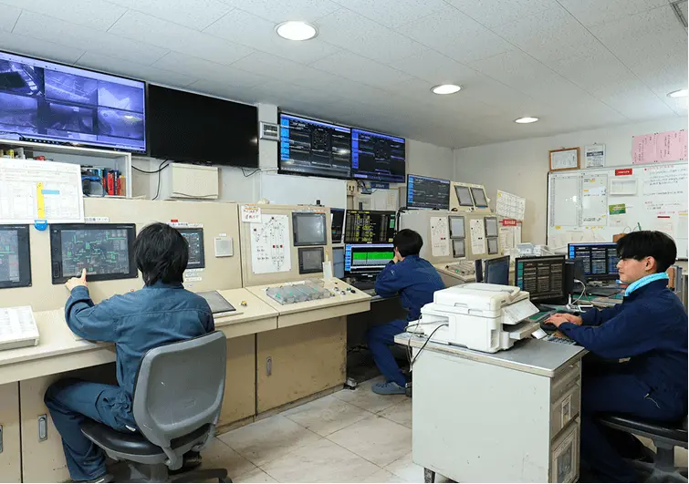 複数の作業員が監視室で大型モニターや制御盤を操作している様子。壁一面の画面に各種データやカメラ映像が映し出され、プラントの運転管理を行っている。