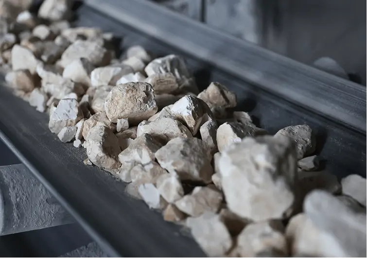 コンベアの上に白色から淡いベージュ色の原料石が多数並んで運ばれている様子。採掘された石灰石などの鉱石が加工工程に送られている。