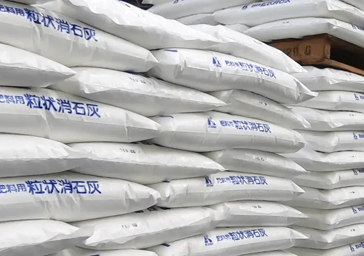 「「肥料用 粒状消石灰」と印字された白い袋が積み重ねられて保管されている様子。古手川産業の粒状消石灰製品で、農業用肥料や土壌改良に使用される製品の出荷・保管風景。