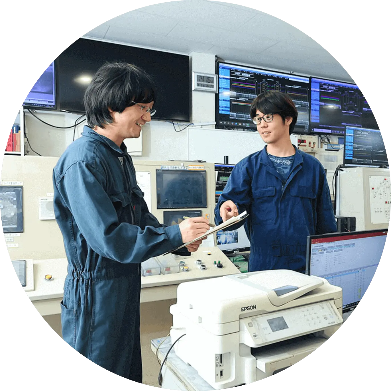 操作室で作業員が機器の前に立ち、資料を手に話し合っている様子
