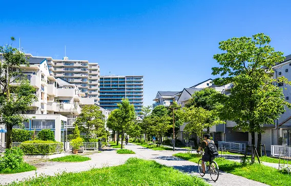 晴天のもと緑豊かな遊歩道を自転車で走る人と、周囲に並ぶ住宅街の風景