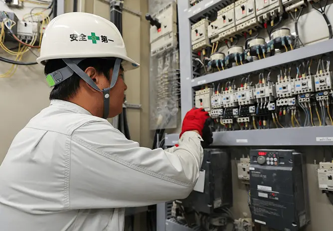 配電盤の前で作業員がヘルメットと手袋を着用し、電気設備の点検作業を行っている様子。多数の配線やスイッチが並ぶ制御盤を慎重に操作している。