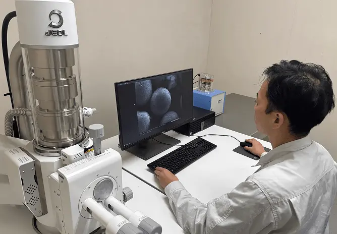 分析装置の前で作業員がコンピューター画面に表示された粒子の拡大画像を確認している様子。電子顕微鏡を用いて材料の表面状態を観察・分析している場面。