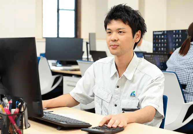 オフィスでパソコンを操作する社員の男性。作業服を着用し、明るい職場環境の中でデスクワークを行っている様子。