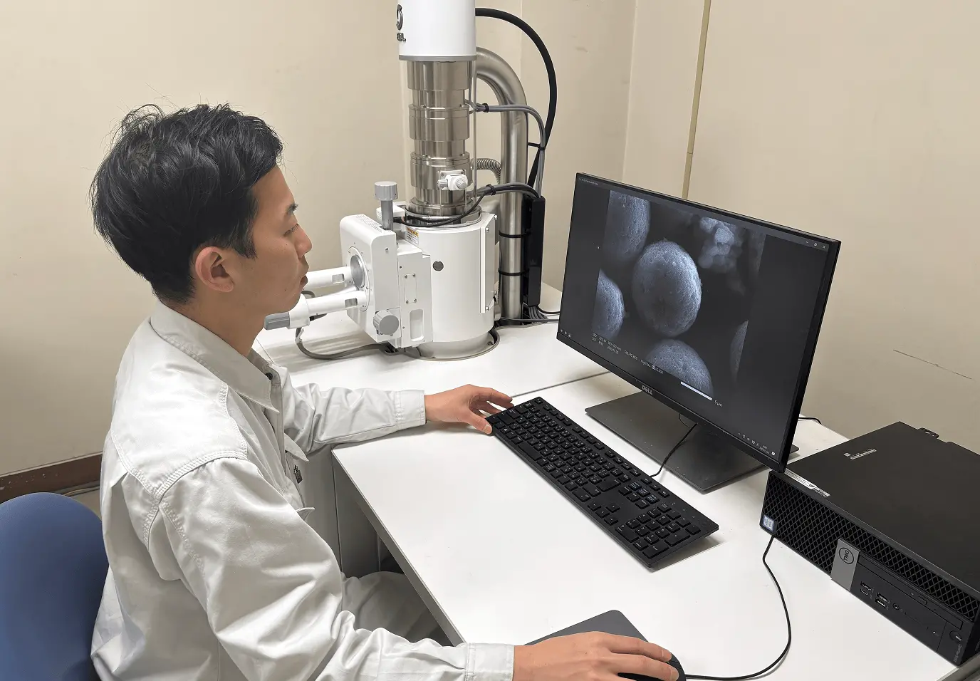 オフィスのデスクで固定電話を使って通話しながら、パソコンのマウスを操作している作業服姿の若手社員。デスクには書類とカレンダーが置かれている