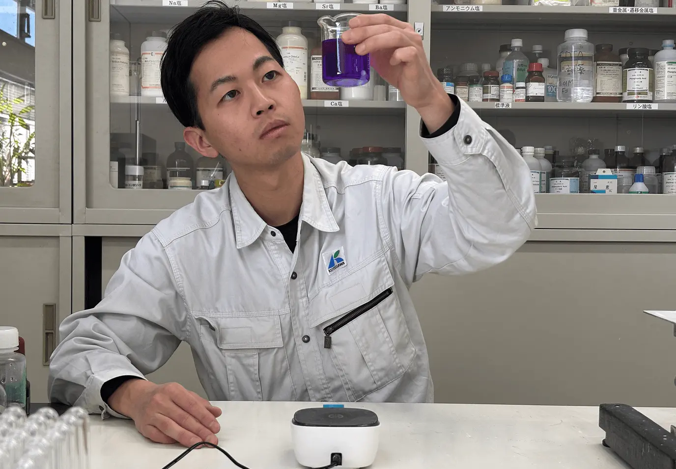 工場内で、白いヘルメットと防塵マスクを着用した作業員が、計測機器を使って「水」と書かれた大型のフレコンバッグ(袋)を検査している様子。背景には天井から吊り下げられたクレーンフックが見える