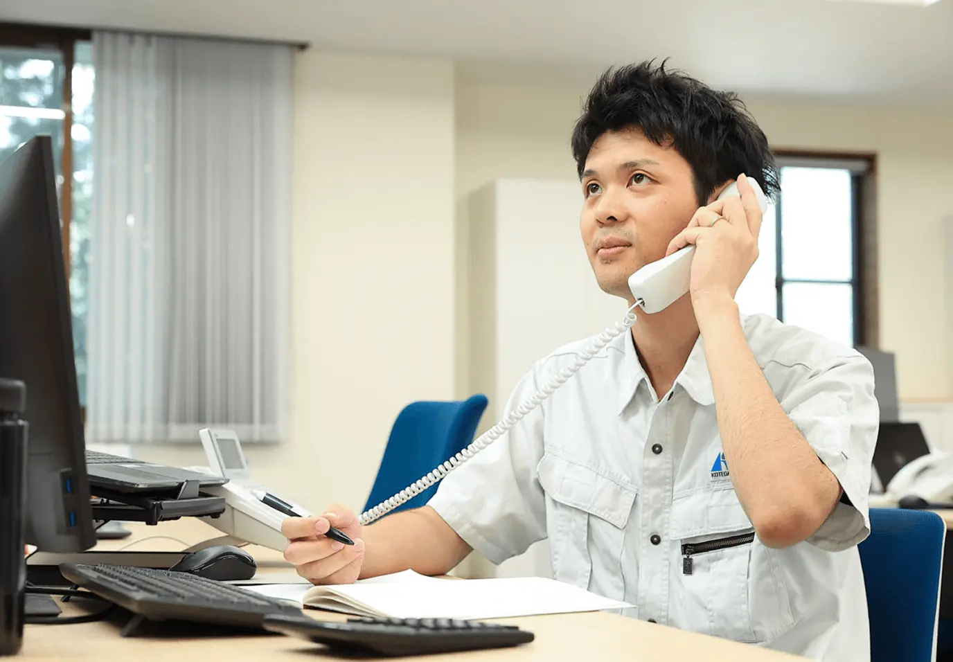 白い作業服を着た男性がオフィスでデスクに座り、電話で通話している様子。デスクにはノートパソコンが置かれ、背景には書類が整理されたファイルキャビネットと本棚が見える。明るく清潔なオフィス環境で、笑顔で電話応対をしている