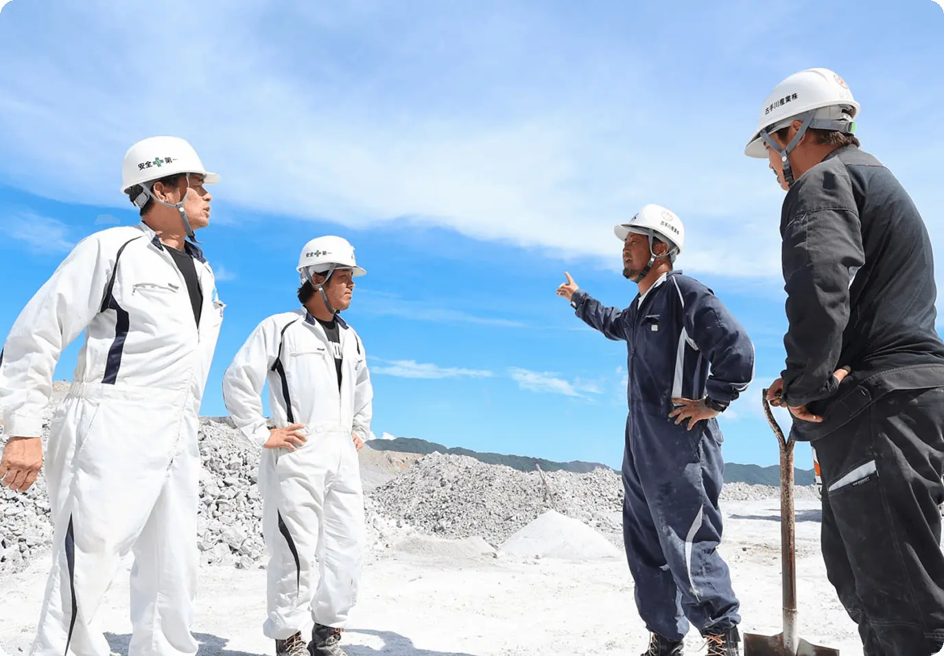 採石場で安全第一と書かれた白いヘルメットを着用した作業員4名が現場で打ち合わせをしている様子。白い作業服と紺色の作業服を着た作業員が、青空の下、白い石灰岩の採掘現場で、1人が指を指しながら説明をしており、他の3名が話を聞いている