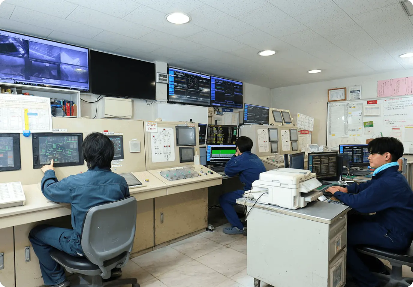 工場の中央制御室で3名の作業員が監視業務を行っている様子。青い作業服を着た作業員たちが、複数のモニターが設置された制御デスクに座り、プラント監視システムを操作している。壁面には監視カメラ映像を表示する大型モニター6台と、プラントの運転状況を示す複数のディスプレイ、各種制御パネルや計器類が配置された24時間体制の監視室