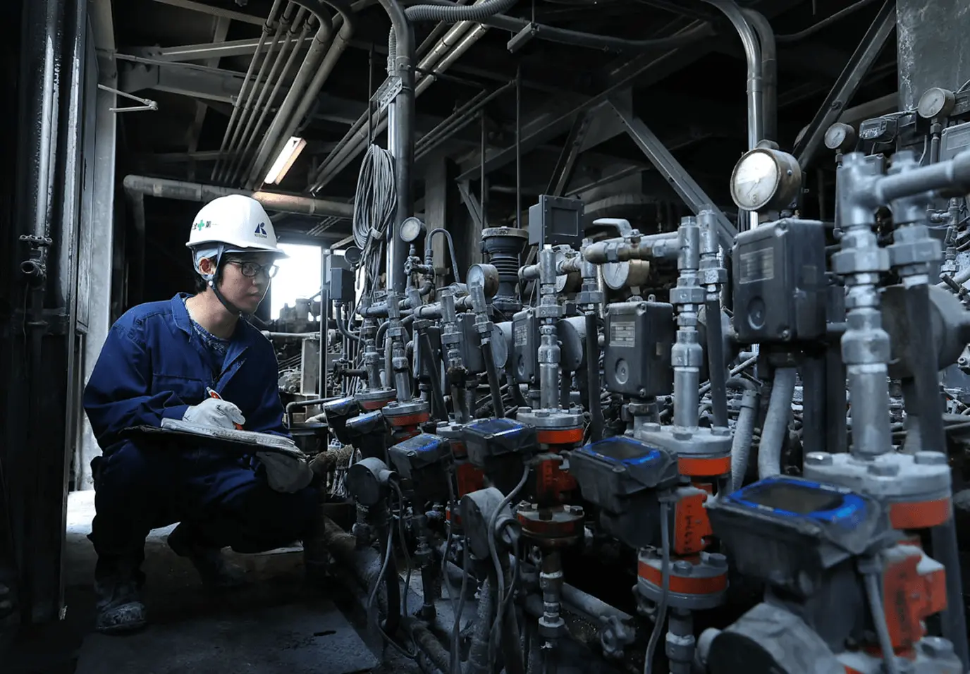 工場内で作業員が安全ヘルメットと保護具を着用し、複数の計器やバルブが並ぶ設備を前に数値を確認しながら点検を行っている様子。配管や計測機器が密集した生産設備の保守作業を示している。
