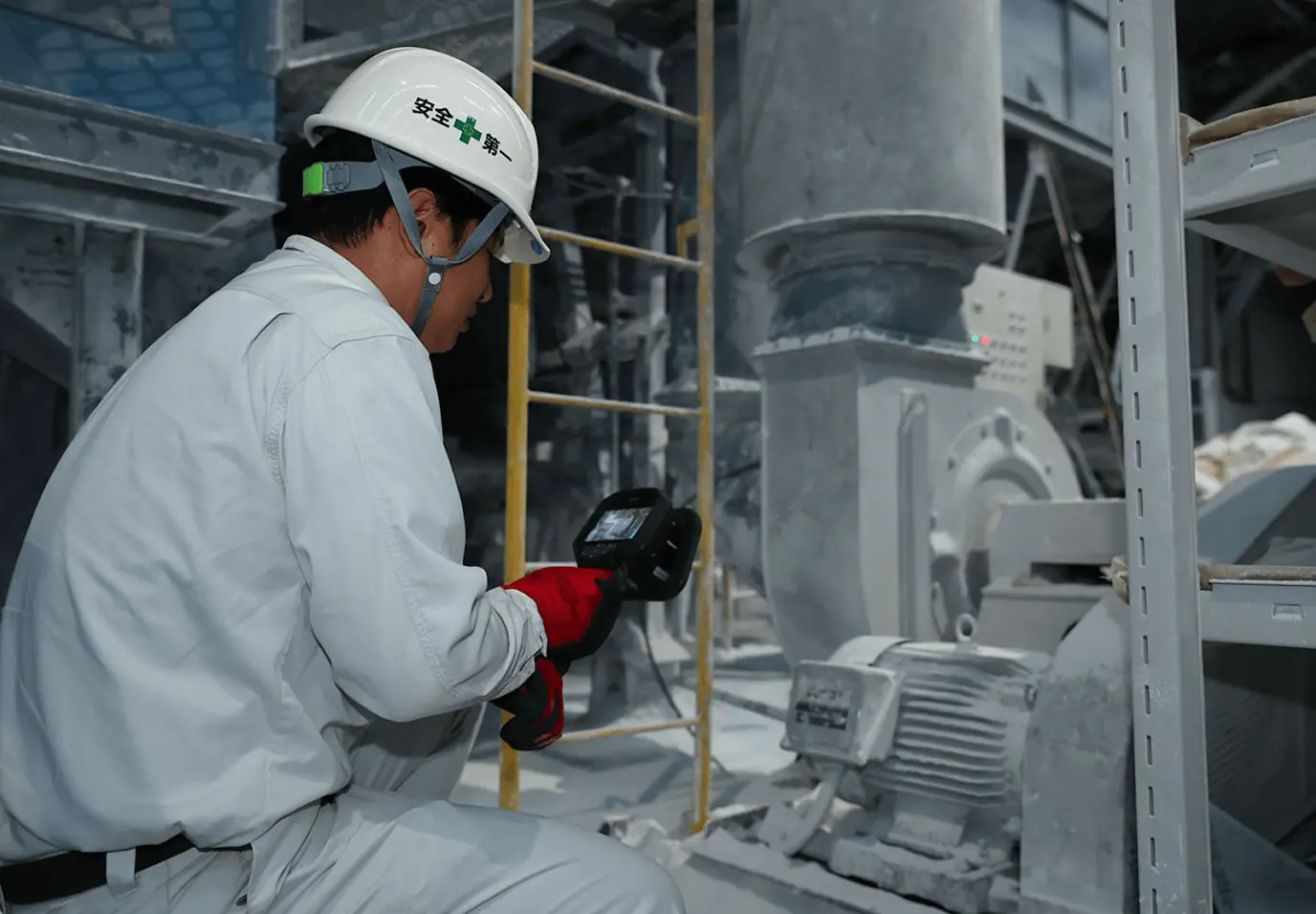 工場内で作業員が安全ヘルメットと手袋を着用し、モーター機器に対してハンディタイプの測定器を使用して点検を行っている様子。粉じんが付着した設備を前に、機器の状態を確認する保守作業を示している。