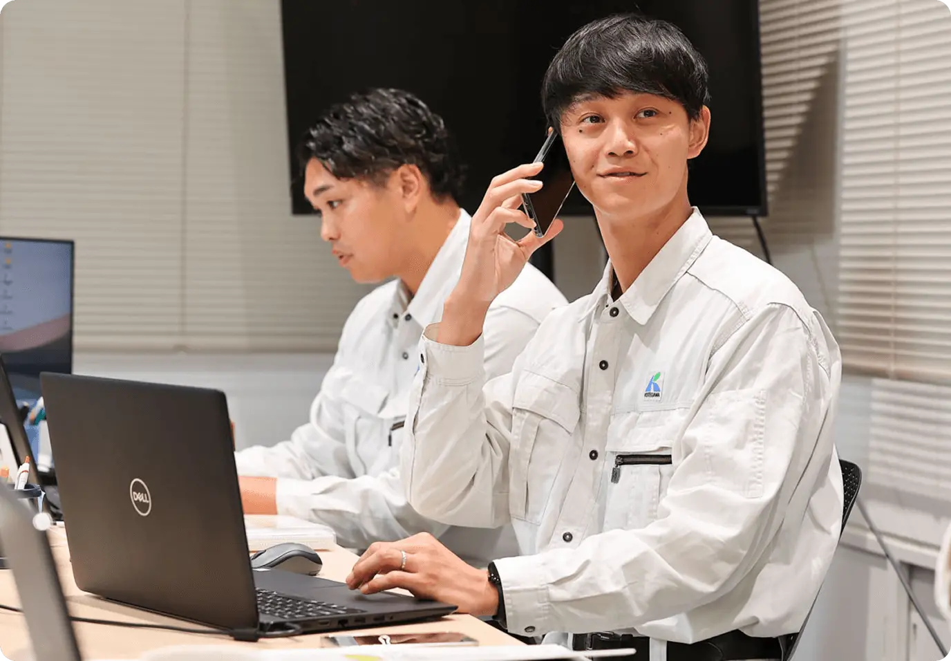 オフィスで2名の営業担当者が業務を行っている様子。手前の男性は白い作業服を着用し、DELLのノートパソコンを操作しながら携帯電話で顧客対応をしている。後ろの同僚もパソコンで作業中。デスクには資料が広げられ、モダンなオフィス環境での営業活動と顧客サポート業務